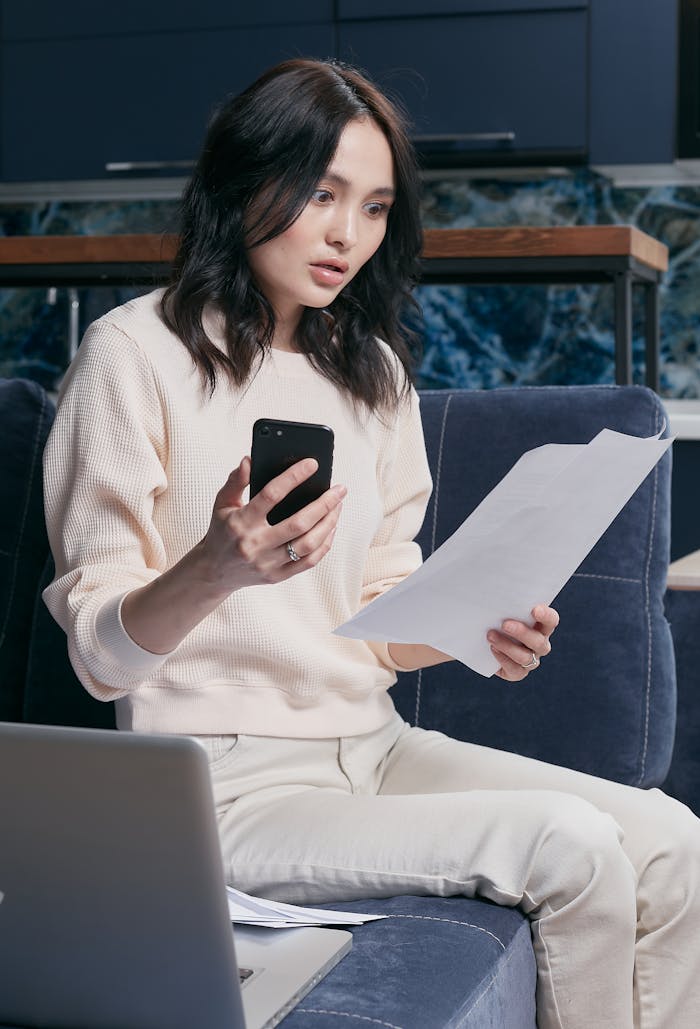 menu-02 A young woman uses her smartphone and laptop to manage finances, appearing focused and concerned.